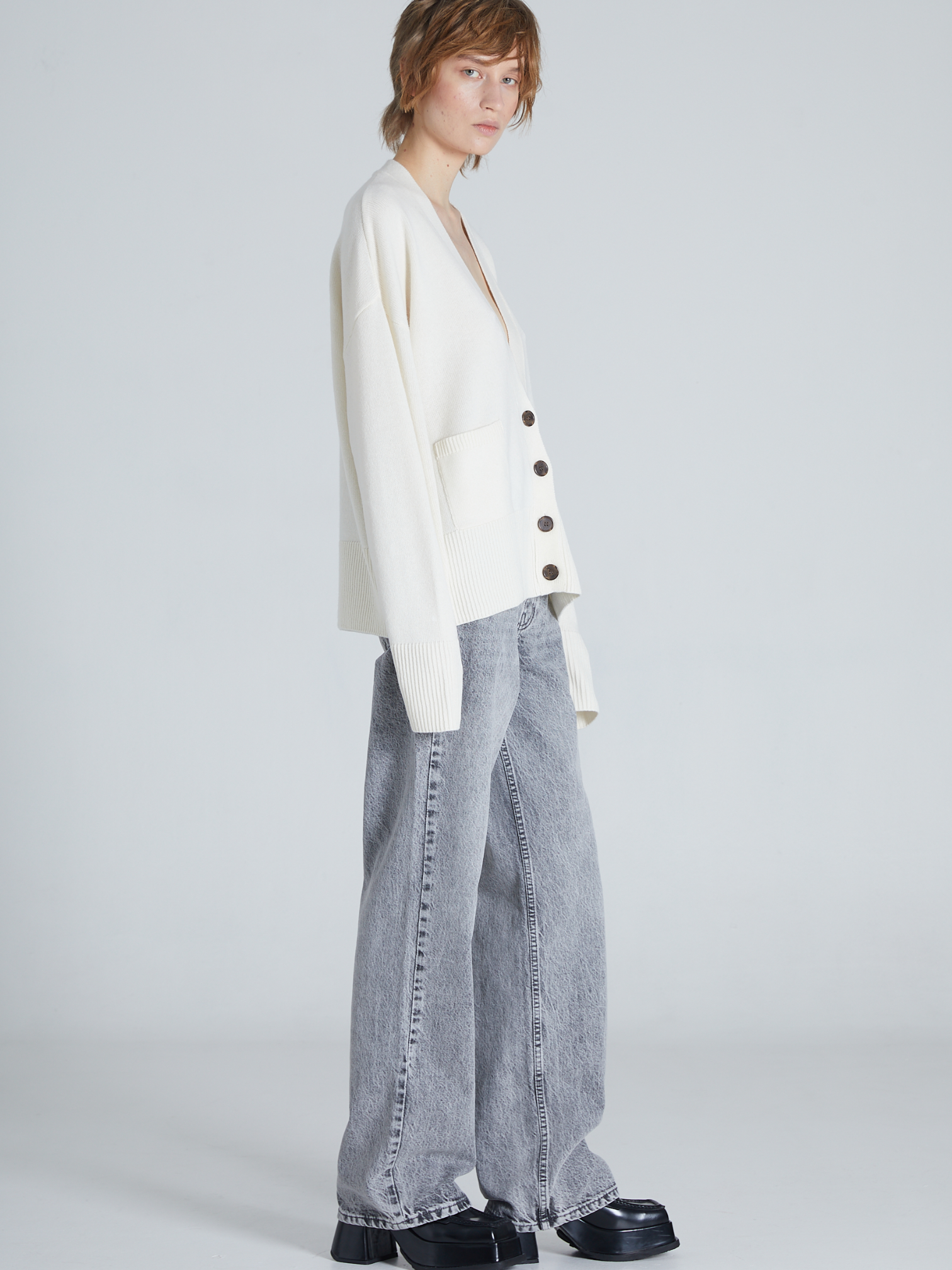 A model is shown from a side profile wearing an oversized off-white cardigan with dark buttons, wide-leg light gray jeans, and black platform shoes, set against a plain light gray studio background.