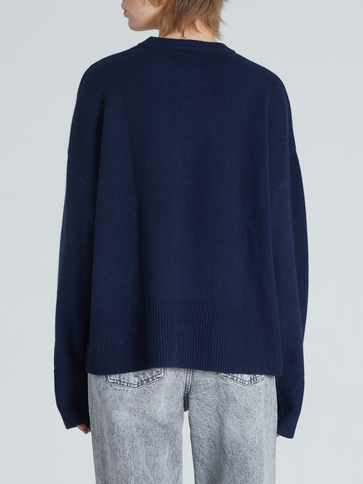 A person shown from the back wearing an oversized navy blue knit sweater and light gray jeans against a plain light gray studio background.