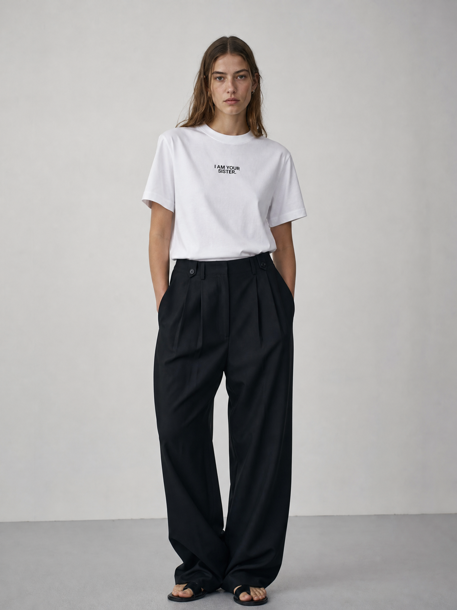 Person wearing a white I am your sister t-shirt and black pants on a plain background
