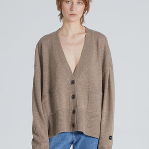 A woman wearing a beige, long-sleeved, V-neck cardigan with front pockets over a plain shirt and blue jeans.