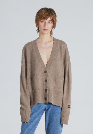 A woman wearing a beige, long-sleeved, V-neck cardigan with front pockets over a plain shirt and blue jeans.