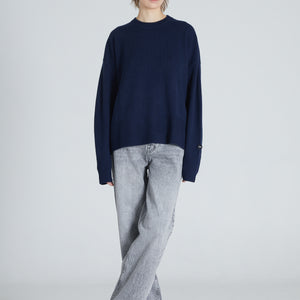 A woman wearing a navy cashmere crew neck sweater and blue jeans, standing against a plain background.