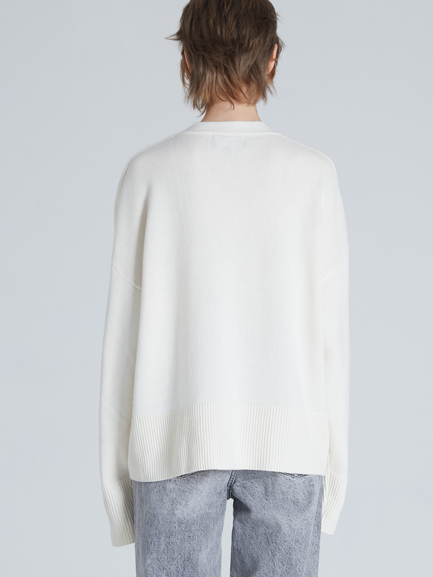A person shown from the back wearing an oversized cream knit sweater and light gray jeans against a plain light gray studio background.