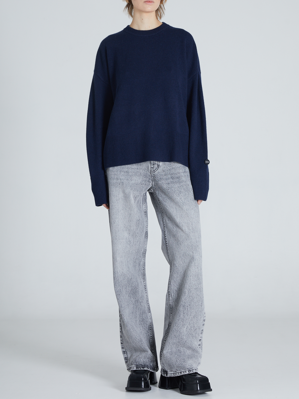 A model wearing a dark navy blue knit sweater and light gray wide-leg jeans, standing against a light gray studio background.