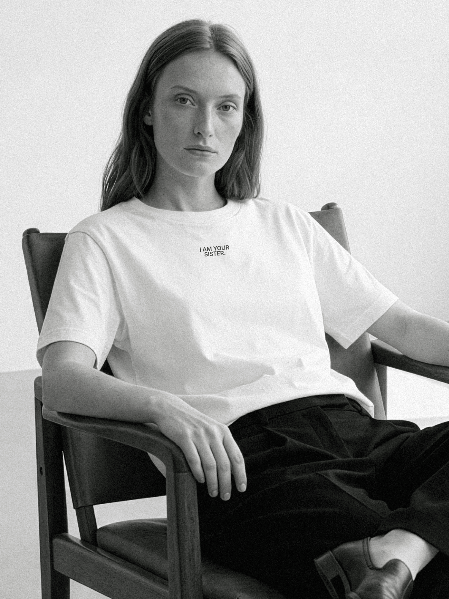 Person wearing a white Sister t-shirt with text, sitting in a chair against a plain background