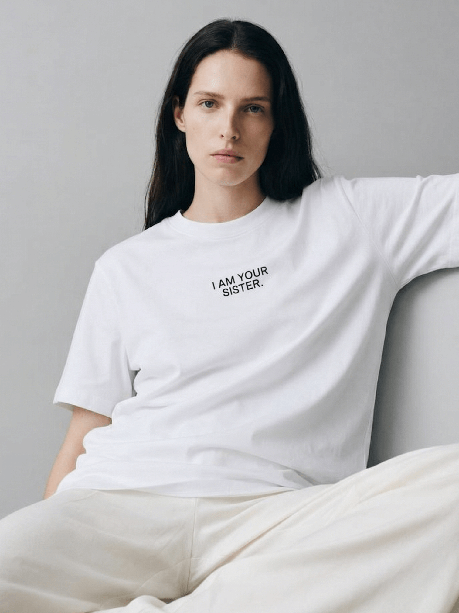 Person wearing a white t-shirt with 'I AM YOUR SISTER' text, sitting on a couch against a plain background.